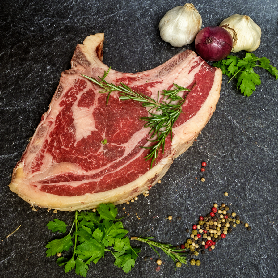 Rib-Eye am Stück mit Knochen und ausgeprägter Marmorierung, mit Rosmarin, Knoblauch und Pfefferkörnern auf dunklem Untergrund.