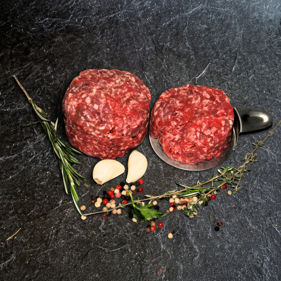 Zwei Portionen Rinder-Hack vom Weiderind, eine davon auf einer Metallschaufel, mit Kräutern, Knoblauch und Pfefferkörnern auf dunklem Steinuntergrund.