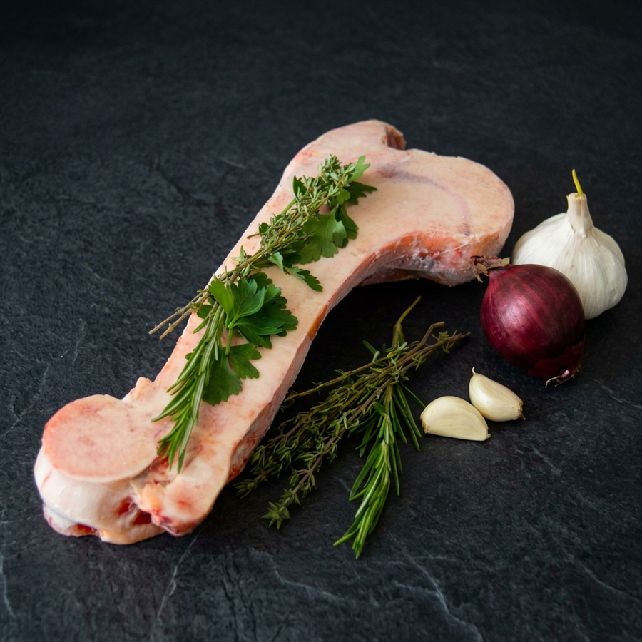 Längs halbierter Markknochen vom Rind mit sichtbarem Knochenmark, Kräutern, Knoblauch und roter Zwiebel auf dunklem Steinuntergrund.