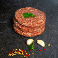 Drei Rindfleisch-Burger-Patties (Beef-Burger) von Gut-Hof-Gruber übereinandergestapelt auf dunkler Steinplatte mit Petersilie, Knoblauch und Pfefferkörnern.
