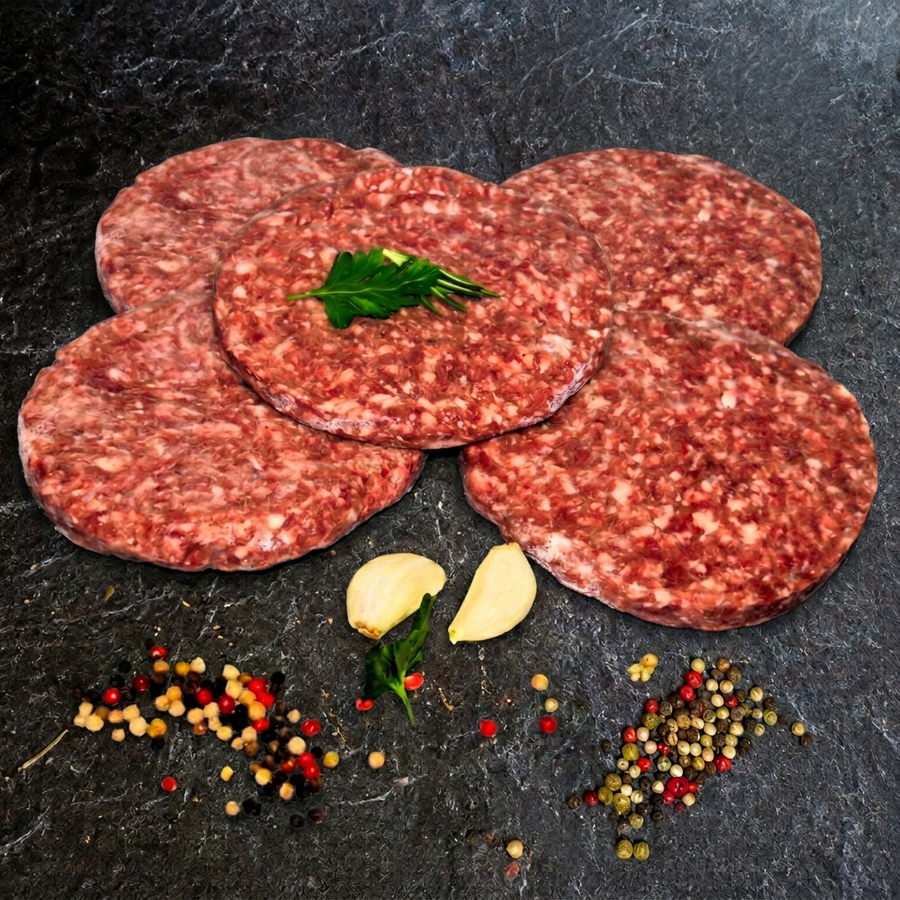 Fünf Rindfleisch-Burger-Patties (Beef-Burger) von Gut-Hof-Gruber überlappend angeordnet mit Kräutern, Knoblauch und Pfefferkörnern auf dunkler Steinplatte.