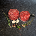 Zwei Portionen Rinder-Hack vom Weiderind, eine davon auf einer Metallschaufel, mit Kräutern, Knoblauch und Pfefferkörnern auf dunklem Steinuntergrund.