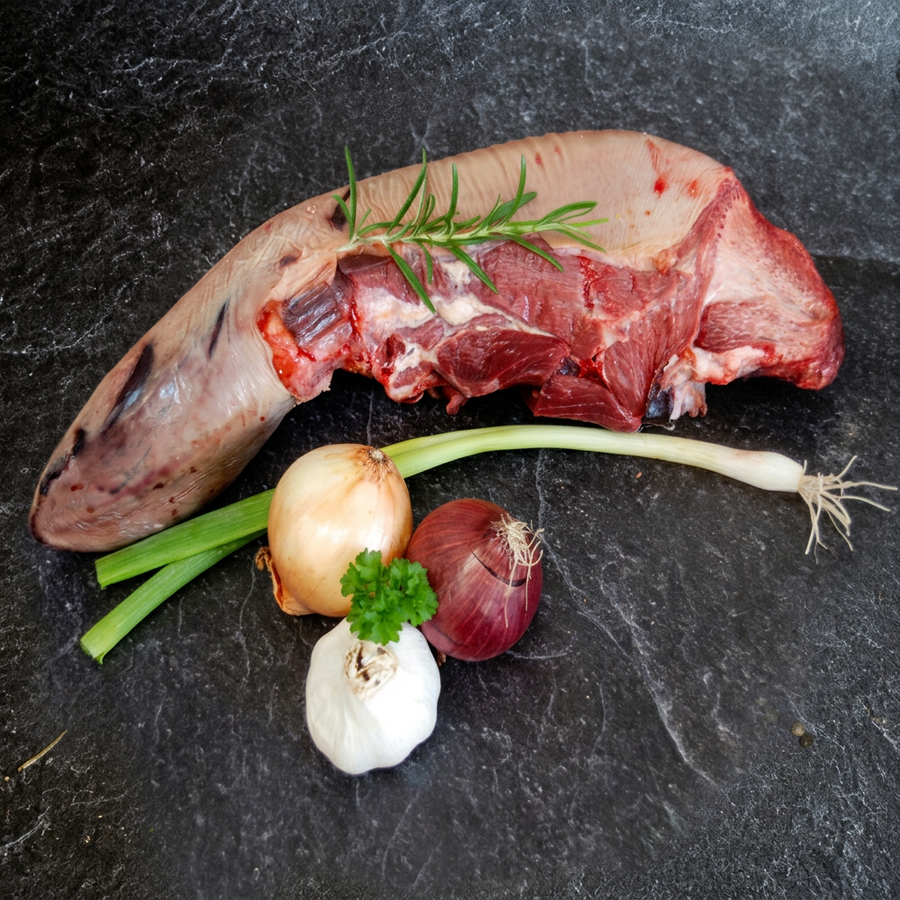 Rinderzunge mit Rosmarin, Petersilie, Zwiebeln, Knoblauch und einer Frühlingszwiebel auf dunklem Steinuntergrund.