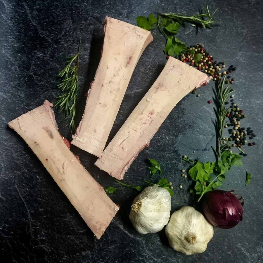 Drei längs gesägte Markknochenstücke vom Rind mit Rosmarin, Petersilie, Pfefferkörnern, Knoblauch und roter Zwiebel auf dunklem Steinuntergrund.